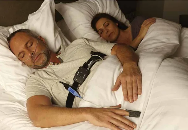 A man sleeping at home with a home sleep apnea test device on his chest and a finger sensor, next to his partner